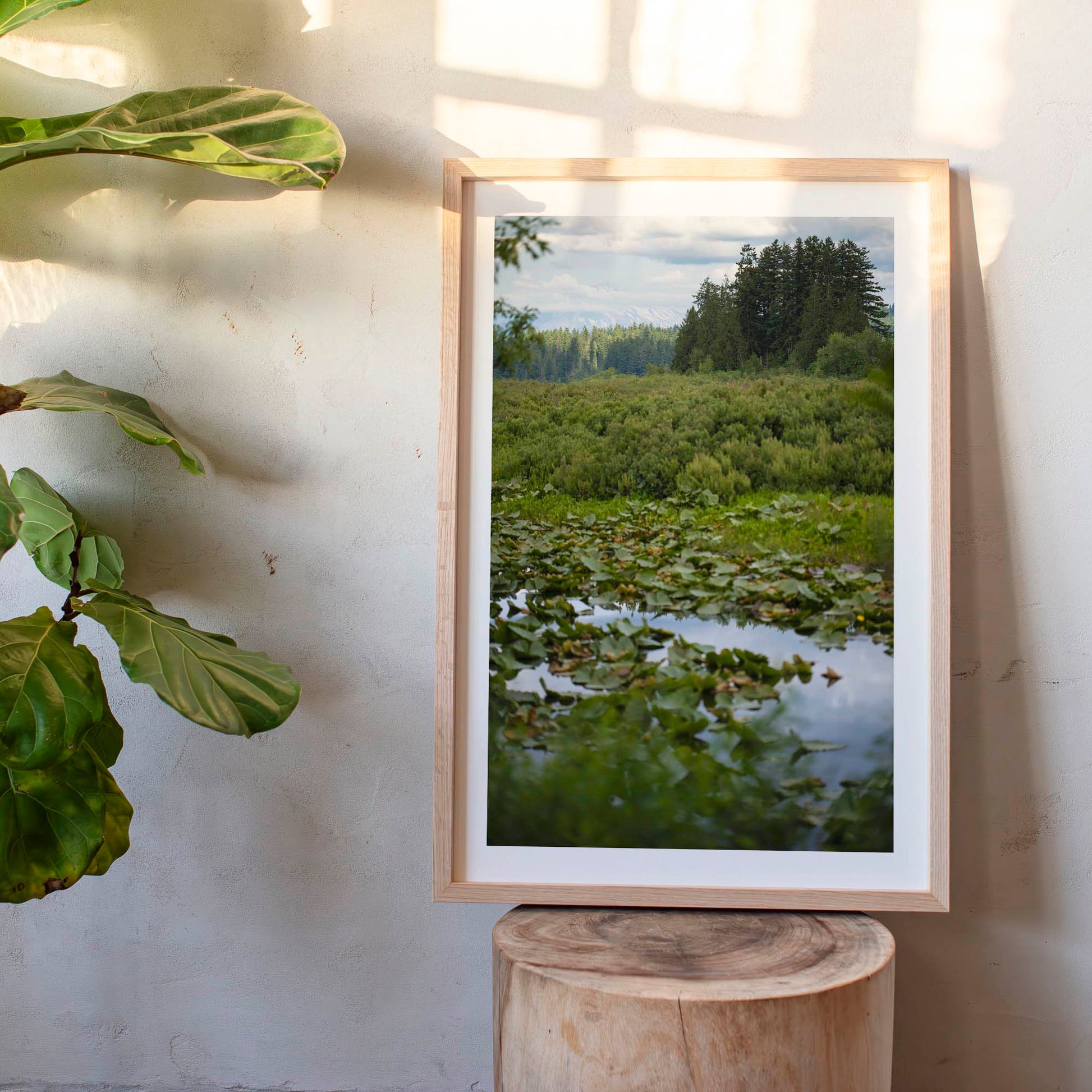 Framed nature print of a forest scene on a wooden stool with a plant to the left.